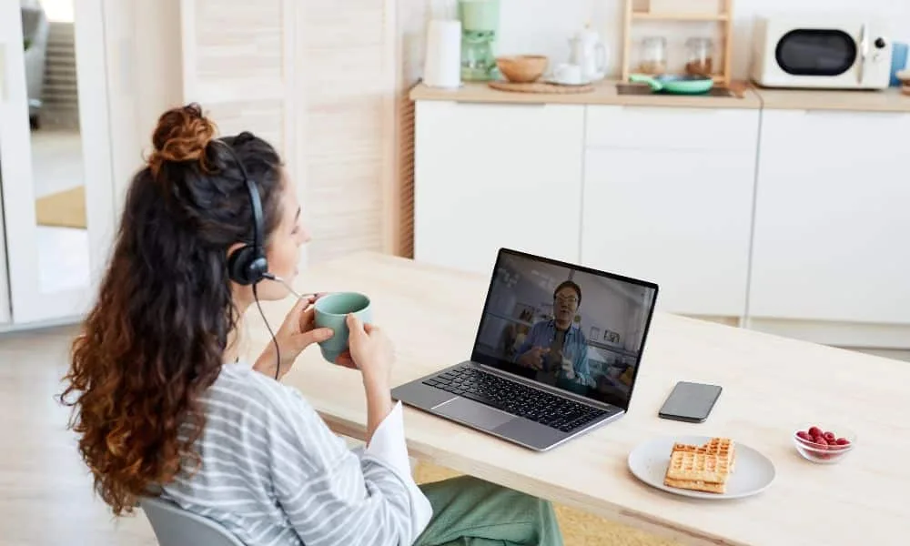 Frau mit dunklen-karamellfarbenen Locken, oberer Teil in einem Topknot, in gestreiftem Hemd, grüner Hose und Headset sitzt am Schreibtisch und führt einen Videocall am Laptop mit Kaffee in der Hand. Waffeln und Himbeeren auf Schreibtisch.
