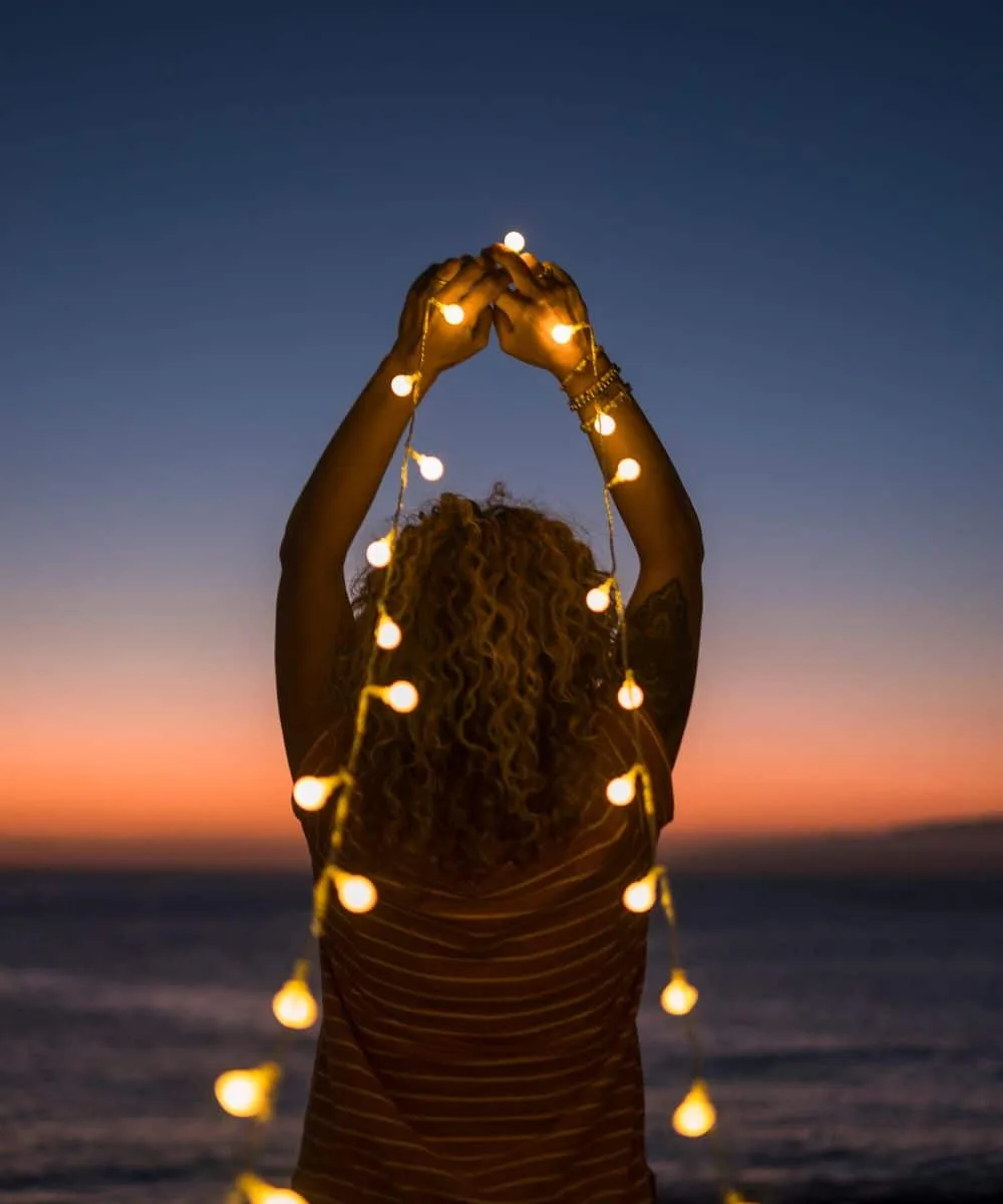 Frau von hinten mit blonden Locken und gestreiftem T-Shirt sitzt in der Abenddämmerung am Meer und hält eine leuchtende Lichterkette hoch.