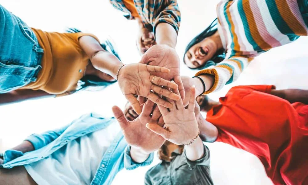 Fünf Menschen legen lachend die Hände in der Mitte zusammen. Weißer Himmel darüber.