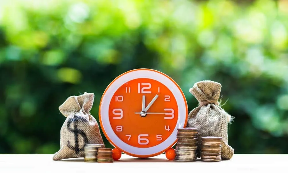 Vier Stapel mit Euro-Münzen und zwei kleine braune Säcke stehen um einen orangenfarbenen Wecker mit weißen Ziffern herum. Grünes Blattwerk im Hintergrund.