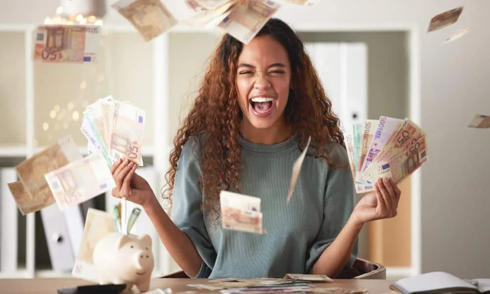 Jubelnde Frau mit rot-braunen Locken, in grauem Shirt am Schreibtisch hat viele verschiedene Geldscheine in der Hand. Andere Geldscheine fliegen durch das Zimmer. Auf dem Schreibtisch ein Sparschwein und ein aufgeklapptes Buch.