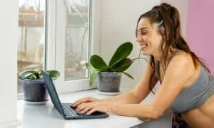 Lächelnde Frau mit dunklem Zopf und grauem Top sitzt am Laptop. Weißer Schreibtisch am Fenster mit Zimmerpflanzen und lila Wand.