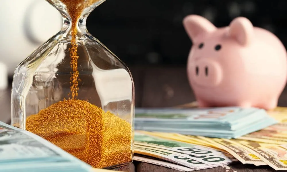 Laufende Sanduhr mit rosa Sparschwein und verschiedenen Geldscheinen auf Tisch. Verschwommener schwarz-brauner Hintergrund.