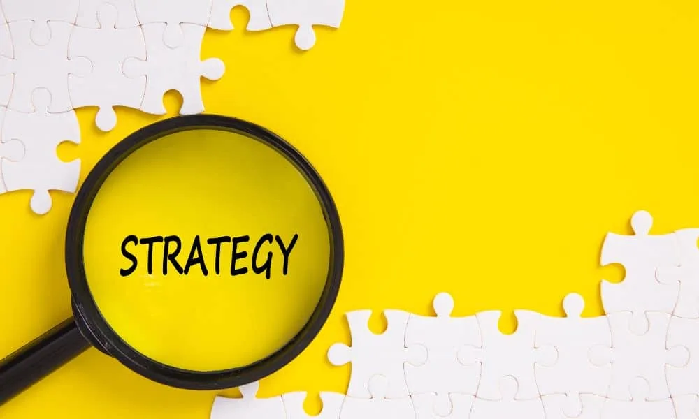 Lupe über dem Wort „strategy“ groß und in schwarz geschrieben, oben und unten zusammengefügte weiße Puzzleteile, gelber Hintergrund.