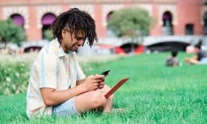 Mann mit braunem Haar und Dreitagebart in gestreiftem T-Shirt und Jeansshorts sitzt lächelnd mit Laptop und Handy im Gras. Ziegelgebäude, Wiese und Menschen verschwommen im Hintergrund.