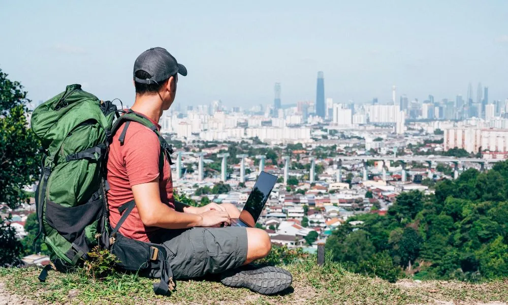 Mann mit dunkler Kappe, rotem T-shirt, grauer Short und grünem Rucksack sitzt mit Laptop auf dem Schoß auf einem Hügel und blickt auf eine Metropole in der Ferne.