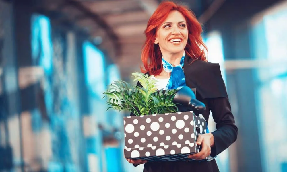 Rothaarige strahlend-lächelnde Frau in dunkler Business-Kleidung und blauem Schal hält einen Karton mit Zimmerpflanze und Tischlampe drin. Verschwommener bläulicher Hintergrund.