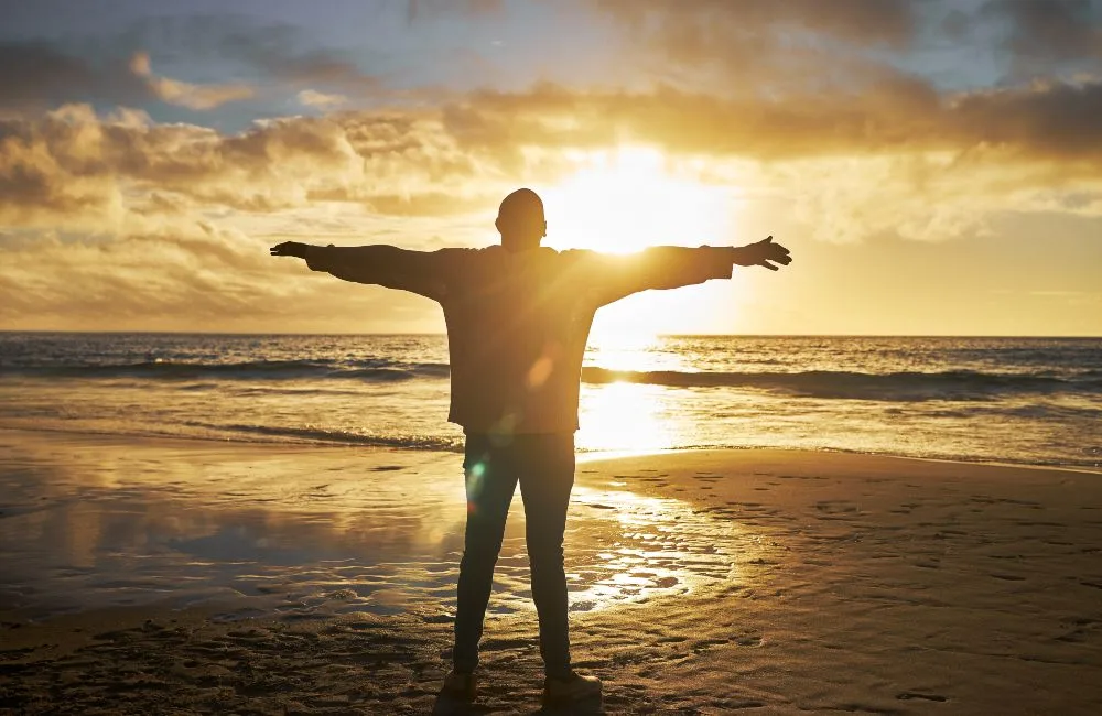 Schattige Silhouette eines Mannes von hinten, die Hände ausgebreitet, steht während eines Sonnenuntergangs am Meer.