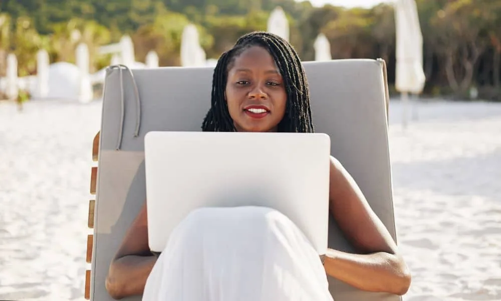 Schwarzhaarige Frau mit rotem Lippenstift und heller Kleidung liegt am Strand auf Liege mit Laptop.