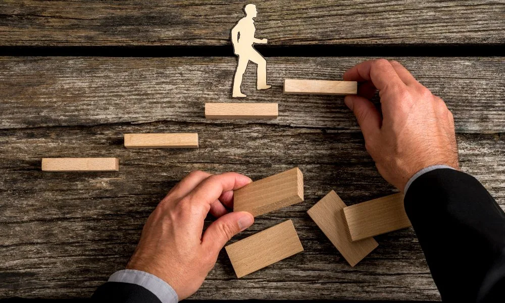 Silhouettenausschnitt eines Mannes, der Stufen hinaufgeht, beides aus hellem Holz, während die Hand eines anderen Geschäftsmannes für ihn Treppen baut. Darunter dunkles Holz.