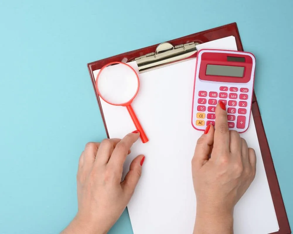 Weibliche Hand mit roten Nägeln drückt auf pinken Taschenrechner, der mit einer roten Lupe auf rotem Klemmbrett liegt. Blauer Hintergrund.