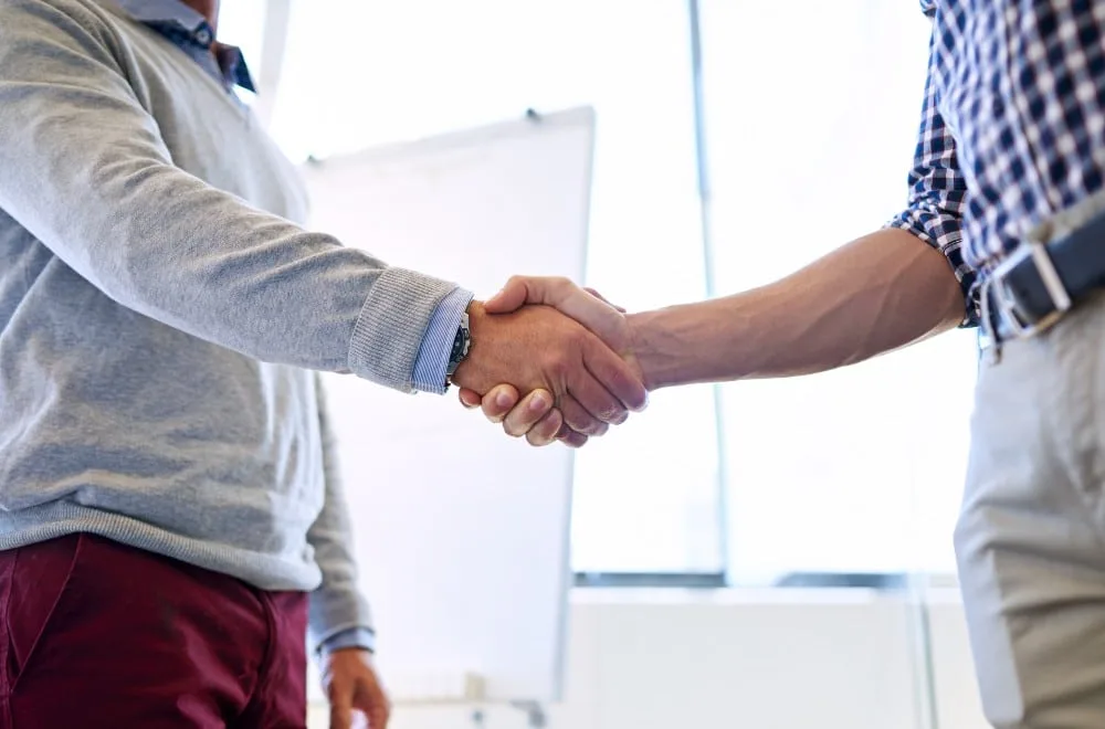 Zwei sich schüttelnde Männerhände, einer im grauen Pullover, der andere im blau-weiß kariertem Hemd. Board und Glaswand dahinter.