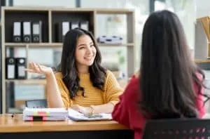 Zwei Frauen sitzen sich am Schreibtisch gegenüber, Regal mit Ordnern im Hintergrund.
