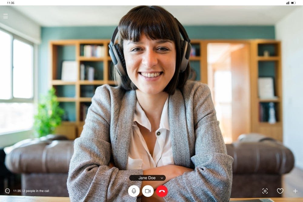 Lächelnde Frau mit dunklem Bob in einem Video Call. Wohnbereich mit blauer Wand, Sofa und Schrank im Hintergrund. 