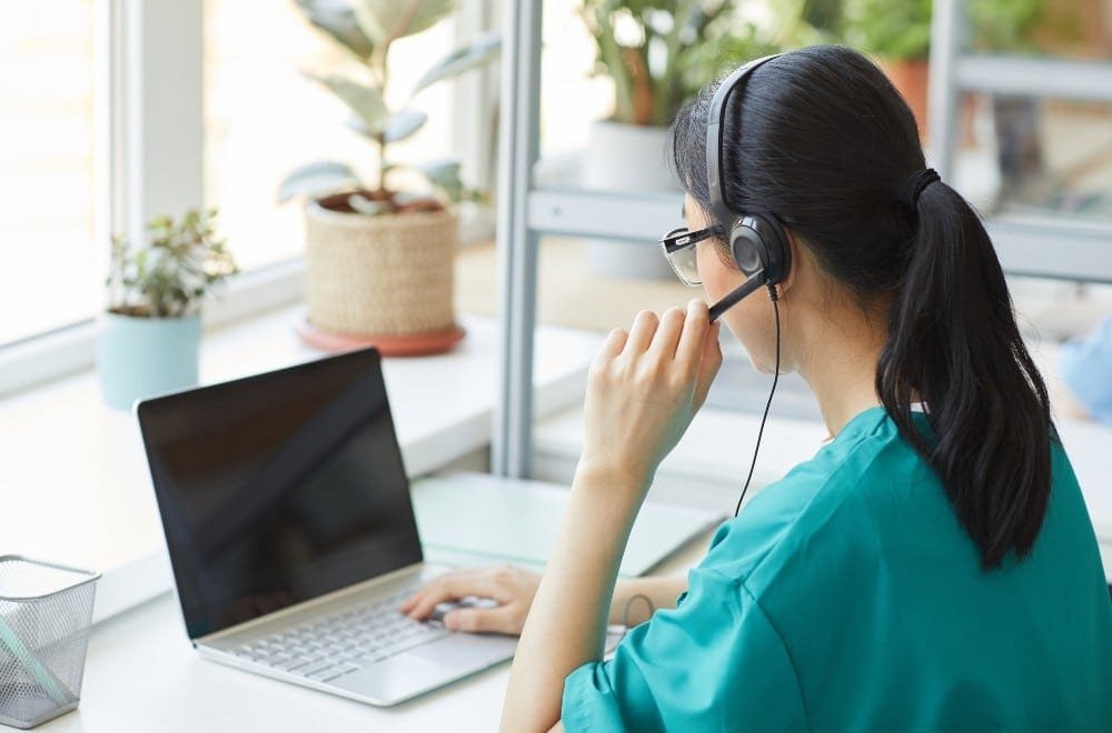 Frau im Profil mit dunklem Zopf, Brille, blauem T-shirt und Headset, die am Laptop sitzt. Verschwommenes Zimmer mit Pflanzen.