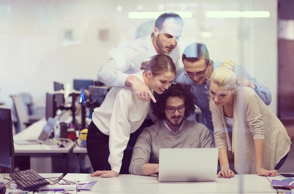 Zwei Frauen und drei Männer stecken die Köpfe zusammen und sehen sich etwas auf dem Laptop an. Hauptsächlich weißer Hintergrund mit Schreibtischen und schwarzen Desktops.
