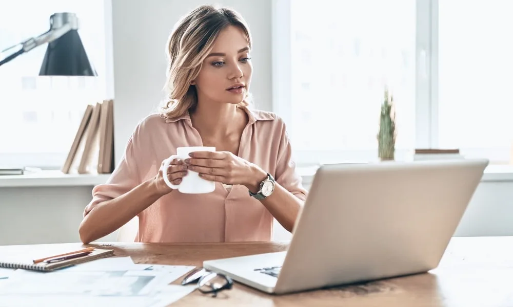 Blonde Frau mit lachsfarbener Bluse und Kaffee in der Hand sitzt vor dem Laptop. Weißer Hintergrund mit Fenstern, Lampe und Zimmerpflanze.