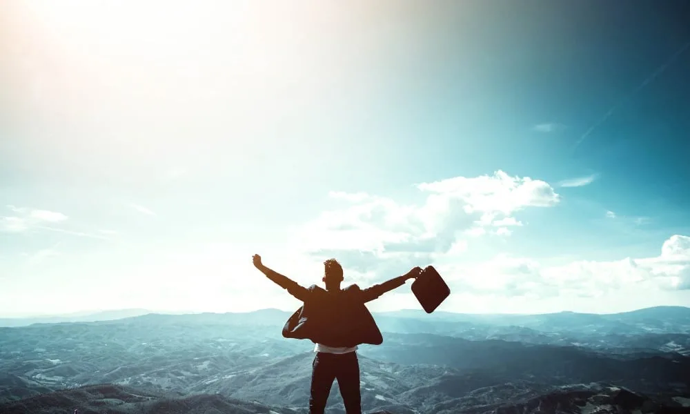 Mann von hinten in dunkler Kleidung und Aktenkoffer in der Hand, steht mit weit offenen Armen vor Gebirge.