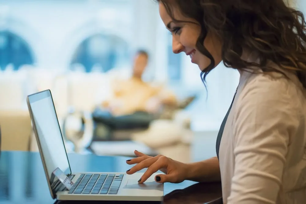 Lächelnde brünette Frau am Laptop im hellen Blazer. Verschwommener Hintergrund mit Mann auf Couch.