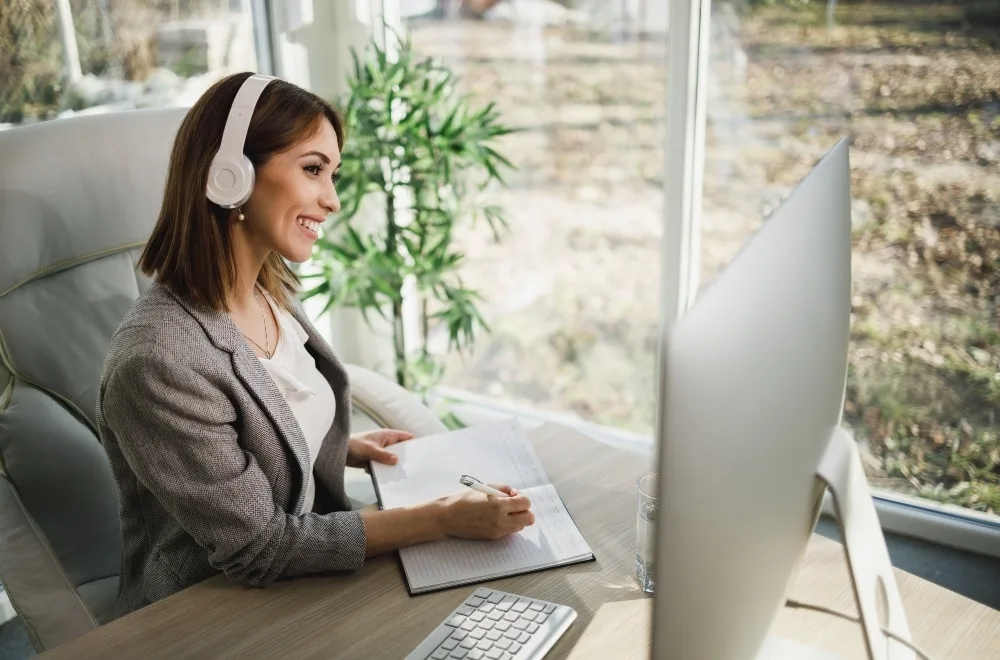 Dunkelblonde, lächelnde Frau in Blazer und Bluse sitzt am Computer mit Headset und schreibt sich was auf. Zimmerpflanze und Terassentür im Hintergrund.