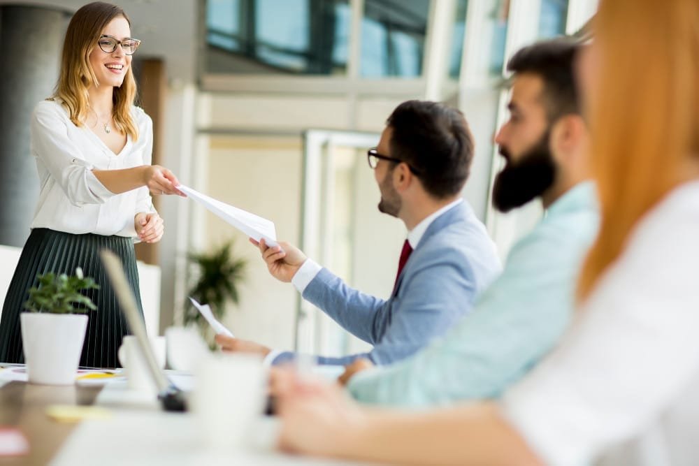 Eine stehende Frau reicht einem sitzenden Mann ein Blatt Papier. Neben ihm sitzen ein weiterer Mann und eine Frau, alle in Businesskleidung in einem Büro.