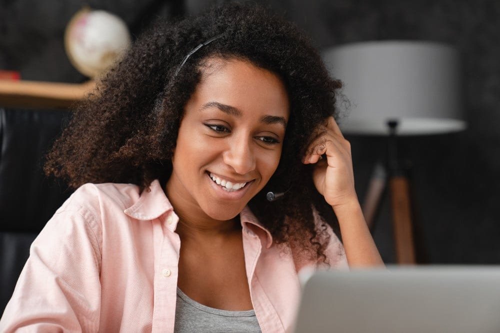 Lächelnde Frau mit dunklen Locken und Headset in rosa Bluse und grauem Top sitzt vor Laptop. Verschwommener Hintergrund mit Lampe und schwarzer Wand.