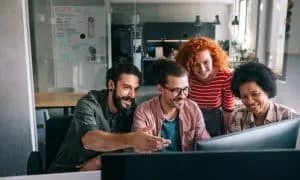Zwei Männer und zwei Frauen sitzen lächelnd vor Schreibtisch mit zwei Bildschirmen. Büro im Hintergrund.