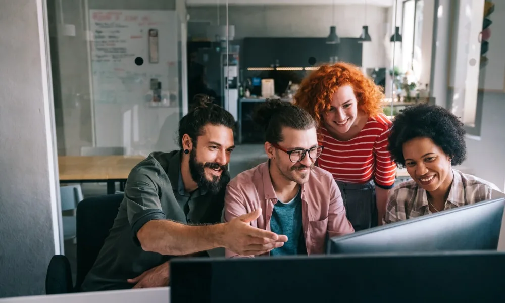 Zwei Männer und zwei Frauen sitzen lächelnd vor Schreibtisch mit zwei Bildschirmen. Büro im Hintergrund.
