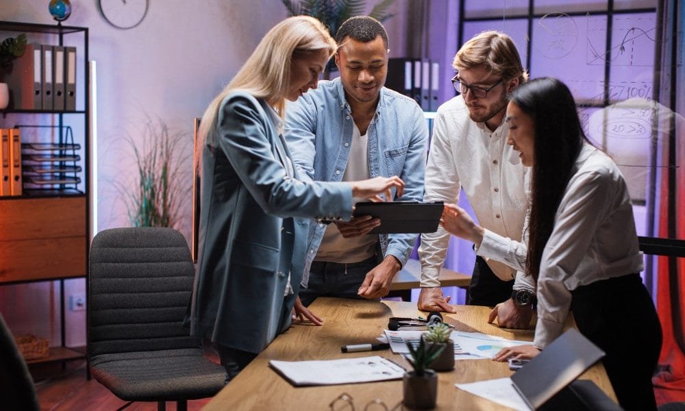 Zwei Männer und zwei Frauen in Businesskleidung stehen um braunen Schreibtisch mit Zimmerpflanzen und Büroutensilien und stecken die Köpfe vor einem Tablet zusammen. Hintergrund mit weißer Wand, Uhr, schwarzem Rahmenfenster und Aktenschrank. 