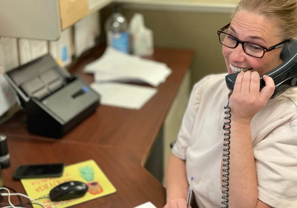 Frau mit blondem Zopf, heller Bluse und Brille telefoniert lächelnd an Schreibtisch.