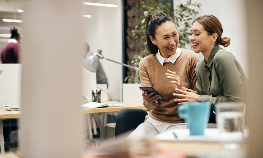 Zwei Frauen, eine im braunen Pullover, die andere in grüner Bluse, sitzen lachend mit Smartphone im Büro. Weißer Hintergrund mit Zimmerpflanze.