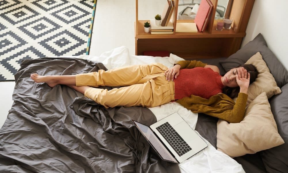 Dunkelhaarige Frau in grünem Cardigan, rotem Top und beigefarbener Hose liegt im Bett mit Hand auf Stirn als Zeichen der Erschöpfung. Laptop mit im Bett.