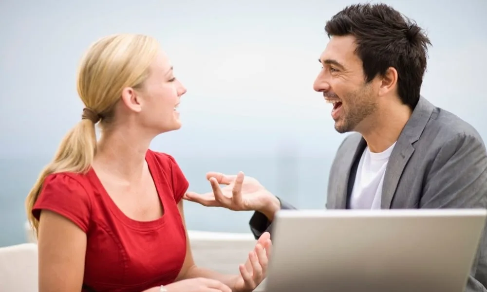 Eine Frau mit blondem Zopf in rotem T-Shirt auf Sofa neben einem dunkelhaarigen Mann mit grauem Sakko und weißem T-Shirt mit Laptop in lebhafter Unterhaltung. Bläulich-weißer Hintergrund.