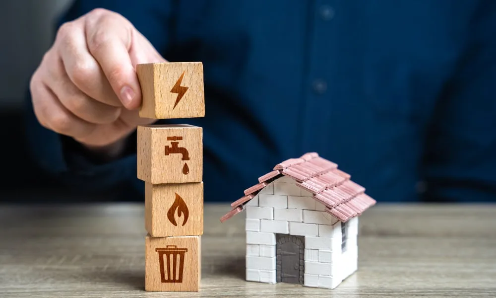 Brustbereich eines Mannes im blauen Anzug sitzt vor Tisch und stellt vier Würfel mit Strom-und-Gas-Abbildungen neben einem Hausmodell auf.