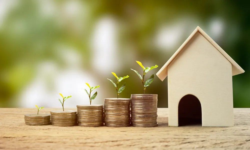 Vier von links nach rechts aufsteigende Münzstapel mit Sprössling drauf enden an einem hellen Holzhausmodell. Darunter helle Holzplatte und verschwommener grüner Hintergrund.