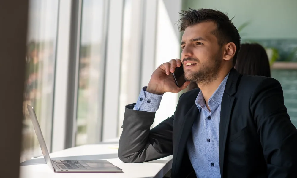 Dunkelblonder Geschäftsmann mit Drei-Tage-Bart und dunklem Anzug sitzt am Laptop und telefoniert am Smartphone. Heller, lichtdurchfluteter Raum mit bodentiefen Fenstern im Hintergrund.