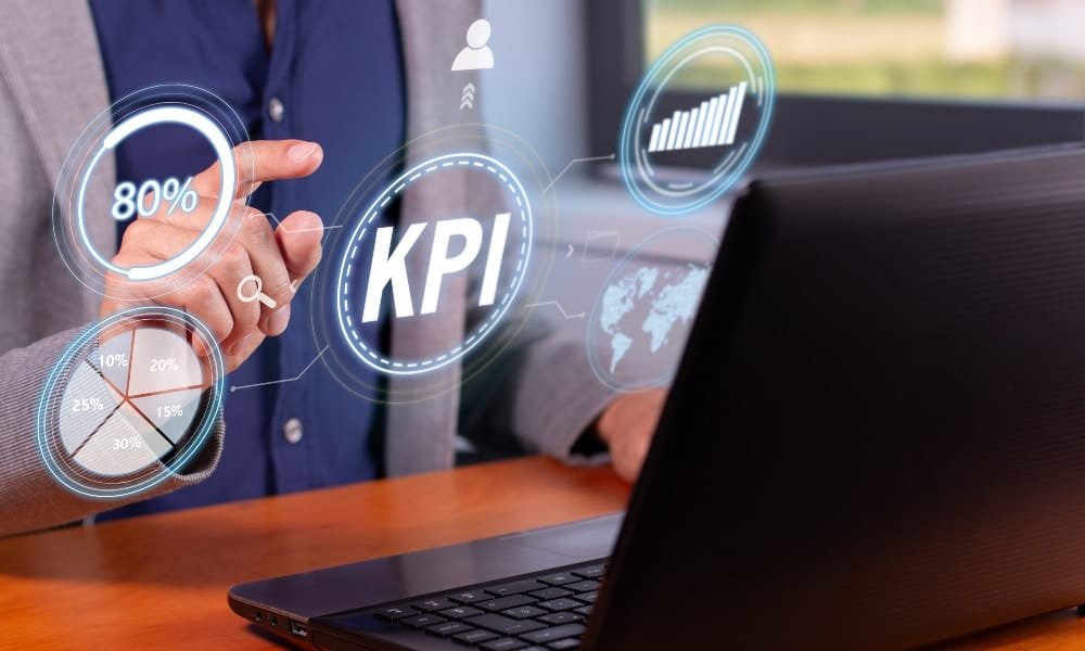 Mann am Schreibtisch mit Laptop vom Brustbereich bis zum Hals in grauem Sakko und blauem Hemd hält eine Hand hoch und zeigt auf das digitale Bild mit KPI, das ein Netzwerk mit anderen digitalen Wabenbildern bildet.