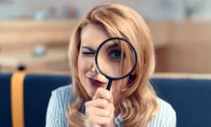 Blonde Frau in weiß-schwarz gestreifter Bluse hält eine Lupe vor das linke Auge und lächelt verschmitzt. Blaue Bank und verschwommener Hintergrund.