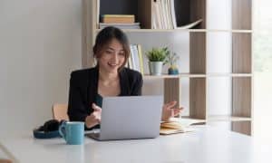 Dunkelhaarige Frau in schwarzem Blazer und grüner Bluse sitzt im lebendigen Gespräch vor Laptop. Weißer Schreibtisch und helles Büro mit Regal im Hintergrund.