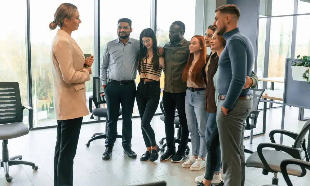 In Büro mit bodentiefen Fenstern stehen sieben Menschen in einer Kette mit Armen um die Schultern oder Taille, und eine blonde Frau davor. Schwarze Schreibtischstühle drumherum. 