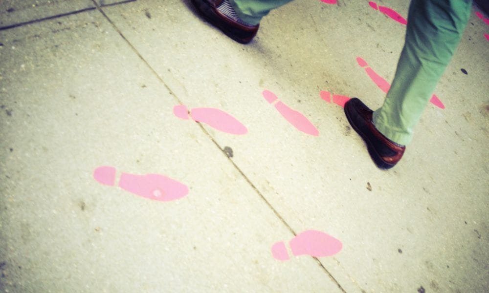 Asphaltweg mit rosa Schuhspuren. Eine Person, von der nur die Beine mit blauer Jeans und schwarz-braunen Schuhen gezeigt wird, geht darüber.