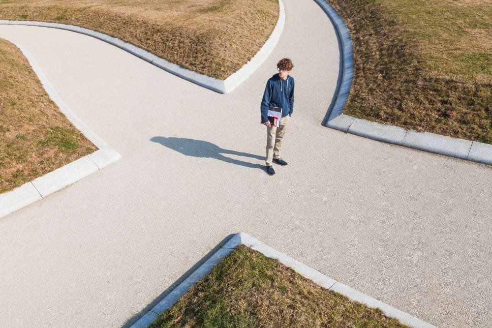 Junger brünetter Mann mit Brille, blauem Hoodie, hellbrauner Hose, blauen Schuhen und Schreibblock unter dem Arm steht inmitten einer Kreuzung aus vier Asphaltwegen und blickt sich um. Trockenes Gras außerhalb der Wege und blauer Himmel im Hintergrund.