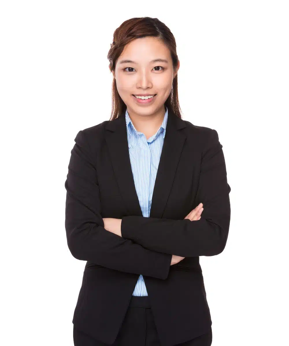 Lächelnde dunkelhaarige Frau mit schwarzem Blazer und hellblauer Bluse steht selbstbewusst mit Armen über Kreuz vor weißem Hintergrund.