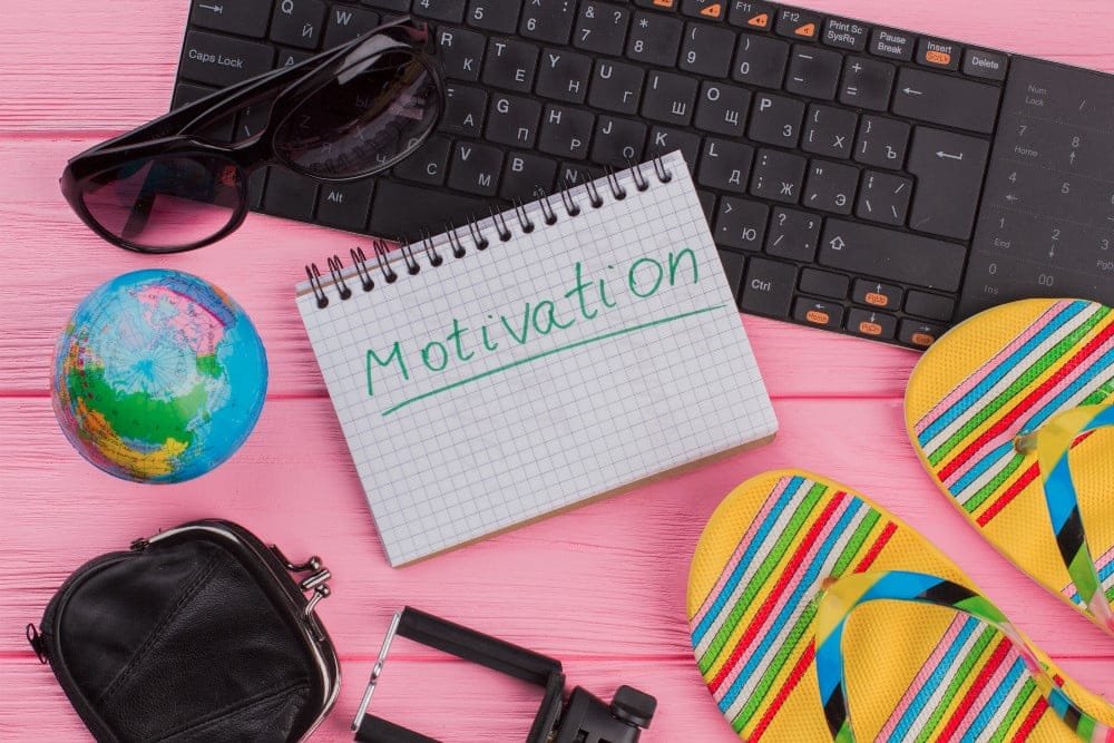Notizblock, auf dem in Grün unterstrichen “Motivation” steht, drumherum Tastatur, Sonnenbrille, Globus, kleine schwarze Tasche und Flip Flops auf pinkem Hintergrund.