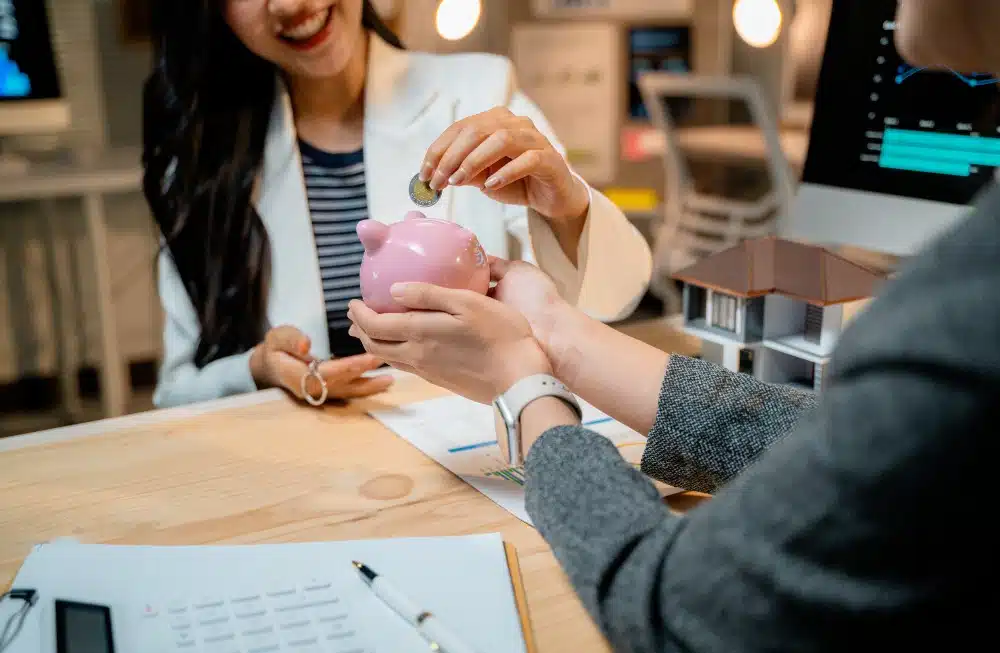 Eine Frau von vorne und eine Frau von hinten sitzen sich am Schreibtisch gegenüber. Eine hält ein rosa Sparschwein hin, die andere wirft eine 2-Euro-Münze hinein. Blatt Papier und Musterhaus auf dem Schreibtisch, verschwommener Hintergrund. 