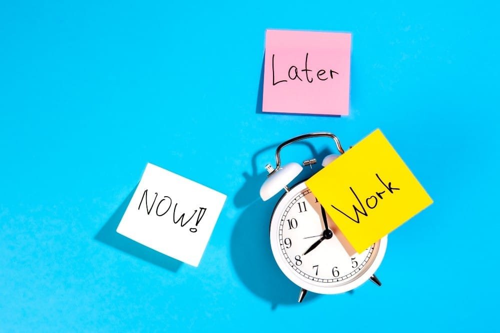 Weißer Wecker auf blauem Hintergrund mit gelbem Zettel, auf dem “Work” steht. Ein rosa Zettel darüber, auf dem “Later” steht, daneben ein weißer Zettel, auf dem “Now” steht. 