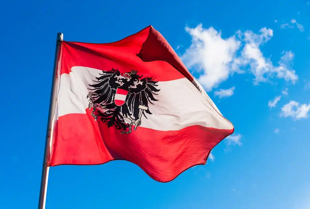 Österreichische Flagge – rot-weiß-rot mit Adler-Wappen in der Mitte – am Mast flattert im Wind, darüber blauer Himmel mit Wolkenfetzen