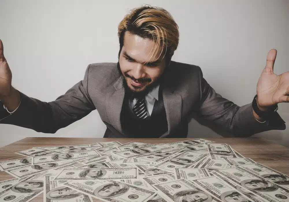 Blond gefärbter Mann mit schwarzem Bart und grauem Anzug ist mit ausgebreiteten Händen lächelnd über Tisch mit einem Haufen Geldscheinen gebeugt. Heller Hintergrund.