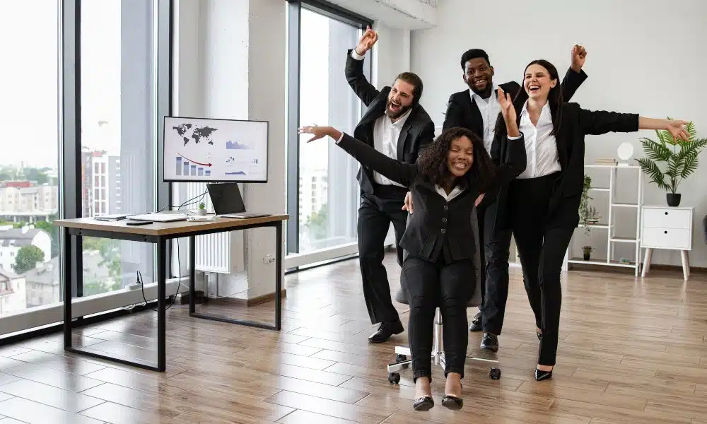 Zwei Frauen und zwei Männer in schwarz-weißer Business-Kleidung stehen zusammen und albern herum in Büro mit bodentiefen Fenstern, weißer Wand mit Regal und Zimmerpflanze im Hintergrund und Schreibtisch mit Computer daneben.