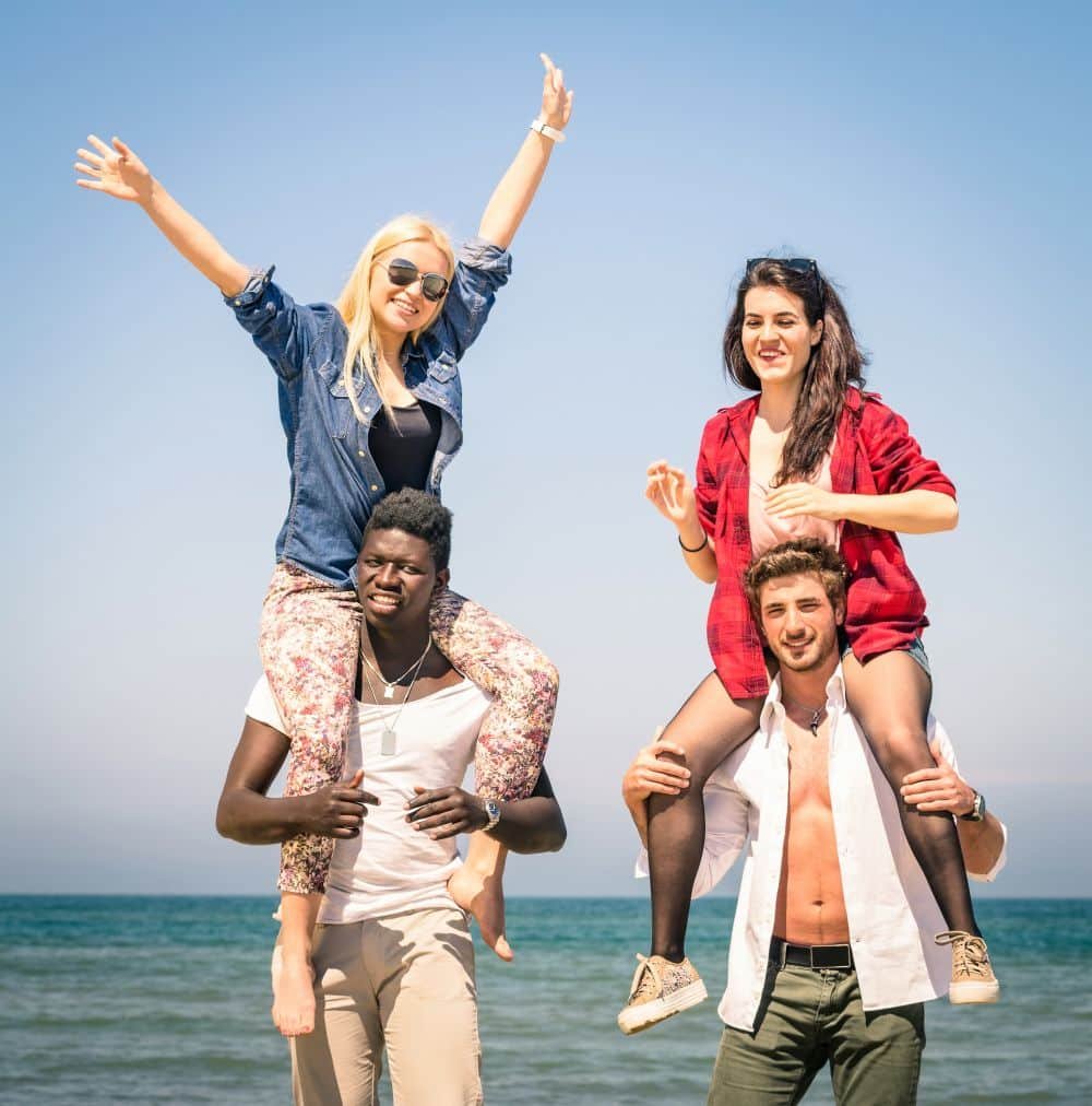 Zwei Männer stehen am Strand und haben beide je eine Frau auf der Schulter, die in die Kamera posieren und Grimassen schneiden.